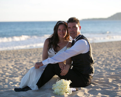 bride and groom sit together on sandy beach