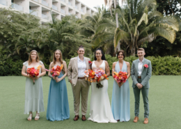 Wedding party posing on a tropical lawn at a Dreams Bahia Mita wedding in Mexico, wearing sage green, blue, and neutral outfits while holding vibrant orange and pink floral bouquets.
