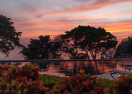 Vibrant pink and orange sunset reflecting across an infinity pool, with tropical trees silhouetted against the sky at Dreams Bahia Mita, a resort near Puerto Vallarta, Mexico.