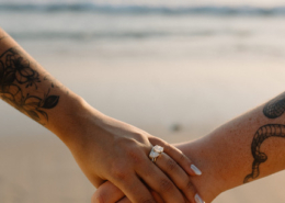 Close-up of two brides holding hands and showing their wedding rings, with floral tattoos visible on their arms and the ocean softly blurred behind them during their Mexico destination wedding.
