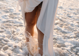 Close-up of a bride lifting her flowing white wedding dress while walking through foamy ocean waves, with floral tattoos visible on her arm during a Puerto Vallarta wedding.