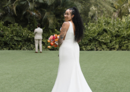 Bride wearing a fitted white wedding gown with a long train, holding a colorful tropical bouquet while looking back toward the camera on a palm-lined lawn at Dreams Bahia Mita.