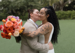 Two brides embracing as one kisses the other's cheek, holding a bright orange and pink tropical wedding bouquet during their romantic Mexico destination wedding celebration.