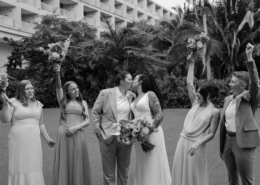 Black-and-white wedding party portrait of two brides kissing while their attendants raise bouquets and cheer on a tropical lawn during their Dreams Bahia Mita wedding in Mexico.