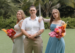 Three wedding attendants posing on a tropical lawn at Dreams Bahia Mita, wearing sage green, beige, and blue outfits while holding vibrant orange and pink floral bouquets.