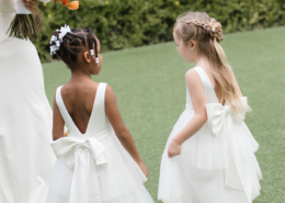 Two young flower girls wearing white tulle dresses with oversized bows and delicate hair accessories, walking together across a green lawn during a Mexico destination wedding.
