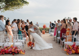 Two brides sharing a romantic dip and kiss while walking down the aisle, surrounded by cheering guests and bright tropical flowers at their Dreams Bahia Mita wedding.