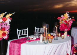 Romantic outdoor wedding reception table decorated with bright pink and orange tropical flowers, glowing candles, and a pink table runner beneath a dramatic Mexico sunset.