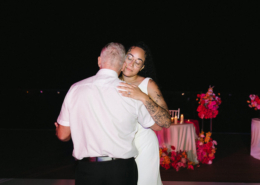 Bride wearing a white wedding gown and glasses sharing a heartfelt dance with an older man during her Mexico destination wedding reception, surrounded by colorful floral arrangements.