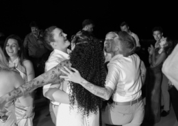 Black-and-white photo of two brides embracing friends and family on the dance floor, surrounded by smiling guests during their Dreams Bahia Mita wedding celebration.