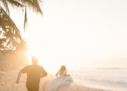 Two brides running barefoot along a sandy beach at sunset, with one lifting her flowing white wedding dress as golden sunlight shines through palm trees during their Dreams Bahia Mita wedding.