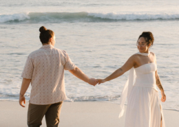 Two brides holding hands while walking along the sandy beach at sunset, one wearing a white dress and the other a patterned shirt, during their Mexico destination wedding celebration.