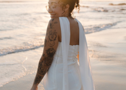 Bride wearing a flowing white wedding dress with long draped fabric, glasses, and floral arm tattoos, smiling over her shoulder on a sandy beach during her Dreams Bahia Mita wedding.