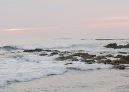 Soft pink sunset over the Pacific Ocean, with gentle waves crashing against a rocky shoreline near Dreams Bahia Mita, creating a peaceful backdrop for a Mexico destination wedding.