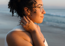 Side-profile portrait of a bride with curly hair, glasses, and a white wedding dress, gently touching her neck as warm golden sunlight illuminates her face beside the ocean in Mexico.