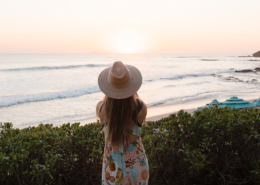 Woman wearing a straw hat and colorful patterned dress looking out over the Pacific Ocean at sunset, surrounded by tropical greenery near Dreams Bahia Mita resort in Mexico.