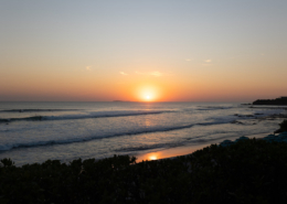 Golden sun setting over the Pacific Ocean near Puerto Vallarta, Mexico, with gentle waves rolling toward the sandy beach and warm orange light reflecting across the water.