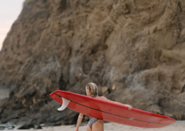 Woman in a swimsuit carrying a long red surfboard along a sandy beach beneath towering rocky cliffs near Puerto Vallarta, Mexico, during a tropical wedding getaway.